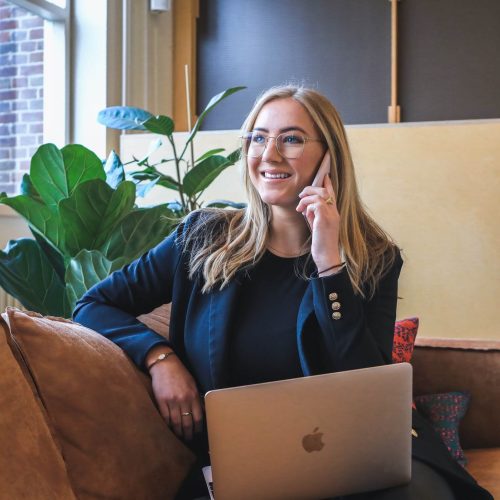 smiling-woman-real-estate-agent-wearing-glasses-sitting-on-sofa-on-phone-with-laptop-building-better-agents